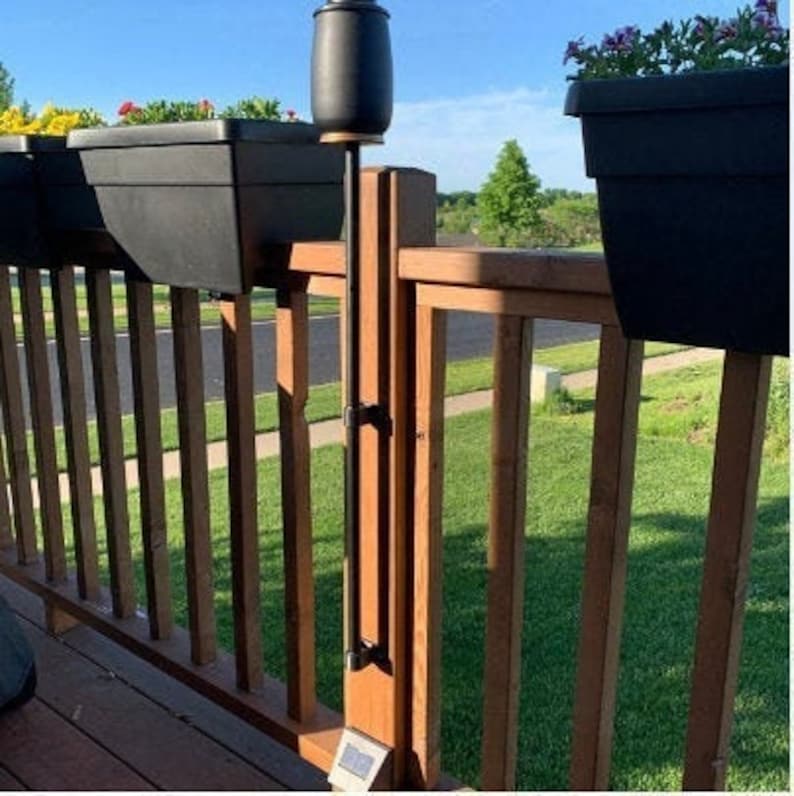 May include: A black metal patio torch holder mounted to a wooden deck railing. The torch holder is attached to a black metal pole that extends from the deck railing to the ground. The torch holder is empty.