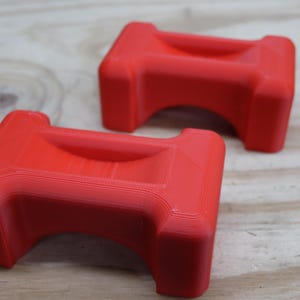 May include: Two bright red, rectangular objects with a curved top and a recessed centre. The objects are placed on a light brown wooden surface. The objects have a smooth, matte finish.