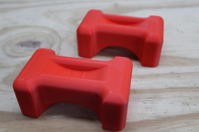 May include: Two bright red, rectangular objects with a curved top and a recessed centre. The objects are placed on a light brown wooden surface. The objects have a smooth, matte finish.