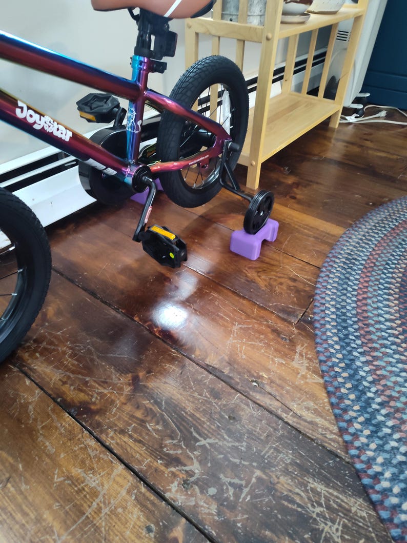 May include: A multi-coloured JoyStar bicycle with training wheels. The bike has a black seat, pedals, and tyres. The training wheels are attached to purple blocks. The bike is on a wooden floor.