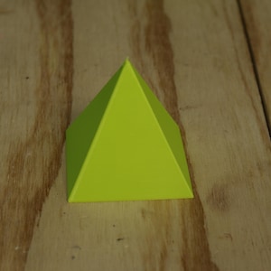 May include: A lime green geometric pyramid shape. The pyramid is resting on a wooden surface. The pyramid has a triangular top and four triangular sides. The object is a solid color and has a smooth surface.