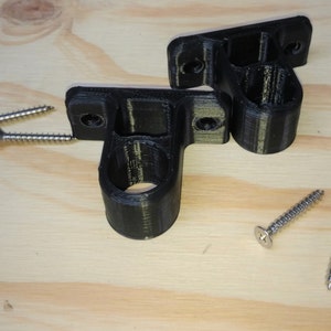 May include: Two black plastic wall mounts with screw holes and a round opening. The mounts are on a light wood surface with two silver screws.