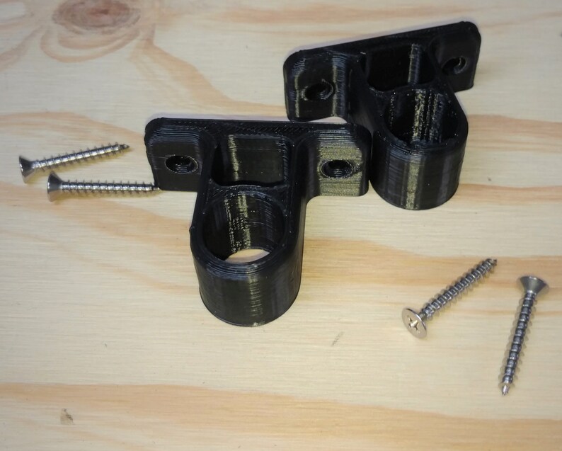 May include: Two black plastic wall mounts with screw holes and a round opening. The mounts are on a light wood surface with two silver screws.