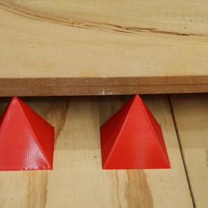 May include: Two bright red pyramid-shaped objects support a piece of wood. The pyramids are on a light-colored wooden surface. The wood above is a darker shade, creating a contrast.