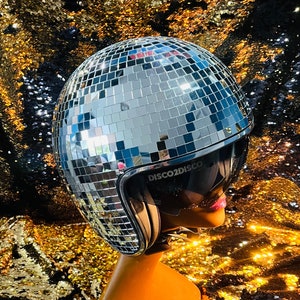 May include: A silver disco ball helmet with a black visor and the text "DISCO2DISCO" is displayed on a mannequin head. The helmet is covered in small, reflective squares. The mannequin head is orange and set against a gold and black sequined background.