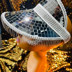 Pode incluir: Uma viseira branca com uma superfície espelhada e uma franja de contas prateadas. A viseira é decorada com strass.