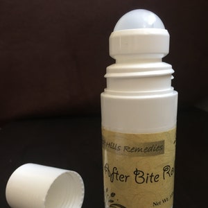 Puede incluir: Botella blanca de roll-on con una etiqueta amarilla que dice "After Bite Roll On" y "Net Wt. 1 fl oz". La etiqueta también tiene un pequeño diseño floral. La botella está sobre una superficie marrón.