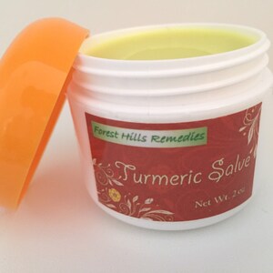 K&ouml;nnte beinhalten: Ein kleines wei&szlig;es Glas mit orangefarbenem Deckel, gef&uuml;llt mit einer gelben Salbe. Das Glas hat ein rotes Etikett mit dem Text "Forest Hills Remedies Turmeric Salve Net Wt. 2 oz."