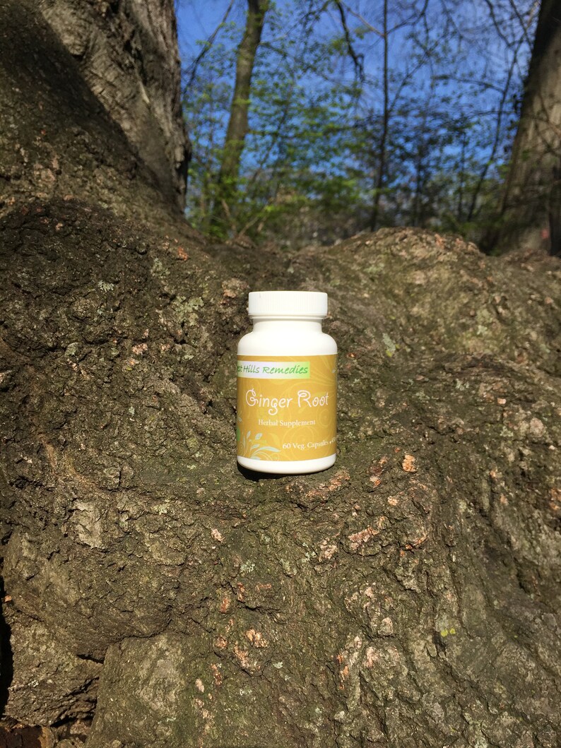 以下が含まれることがあります： グリーンのラベルに「Green Hills Remedies Ginger Root Herbal Supplement 60 Veg Capsules」と書かれた白いボトル。ボトルは木の幹の上に置かれています。