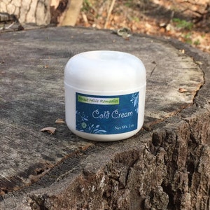 May include: A white jar of Forest Hills Remedies Cold Cream with a blue and green label. The label has a floral design and the text "Cold Cream" and "Net Wt. 2 oz."