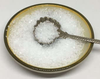 May include: A small, round ceramic bowl with a yellow interior and a green rim, filled with coarse white salt. A decorative silver spoon rests in the salt. The spoon has a scalloped bowl and an ornate handle.