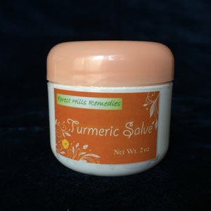 K&ouml;nnte beinhalten: Ein wei&szlig;es Glas mit einem pfirsichfarbenen Deckel und einem Etikett mit der Aufschrift "Forest Hills Remedies Turmeric Salve Net Wt. 2 oz". Das Etikett zeigt eine gelbe Blume und gr&uuml;ne Bl&auml;tter auf orangefarbenem Hintergrund.