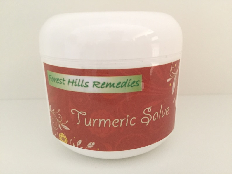 K&ouml;nnte beinhalten: Ein wei&szlig;es Glas mit einem roten Etikett, auf dem "Forest Hills Remedies Turmeric Salve" steht. Das Etikett hat ein florales Design mit gr&uuml;nen Bl&auml;ttern und gelben Blumen.