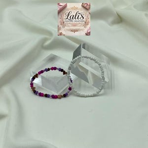 Puede incluir: Dos pulseras, una blanca y otra con cuentas rosas, moradas y doradas. Las pulseras están sobre una superficie blanca.