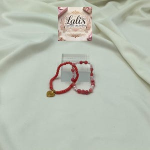 Puede incluir: Dos pulseras de cuentas, una rosa y otra roja, con un dije de corazón dorado. Las pulseras están sobre un fondo blanco.