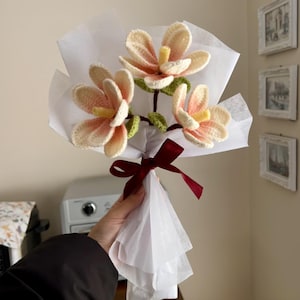 Può includere: Un bouquet di tre fiori di magnolia all'uncinetto in tonalità rosa e bianco, legato con un nastro bordeaux e avvolto in carta velina bianca.