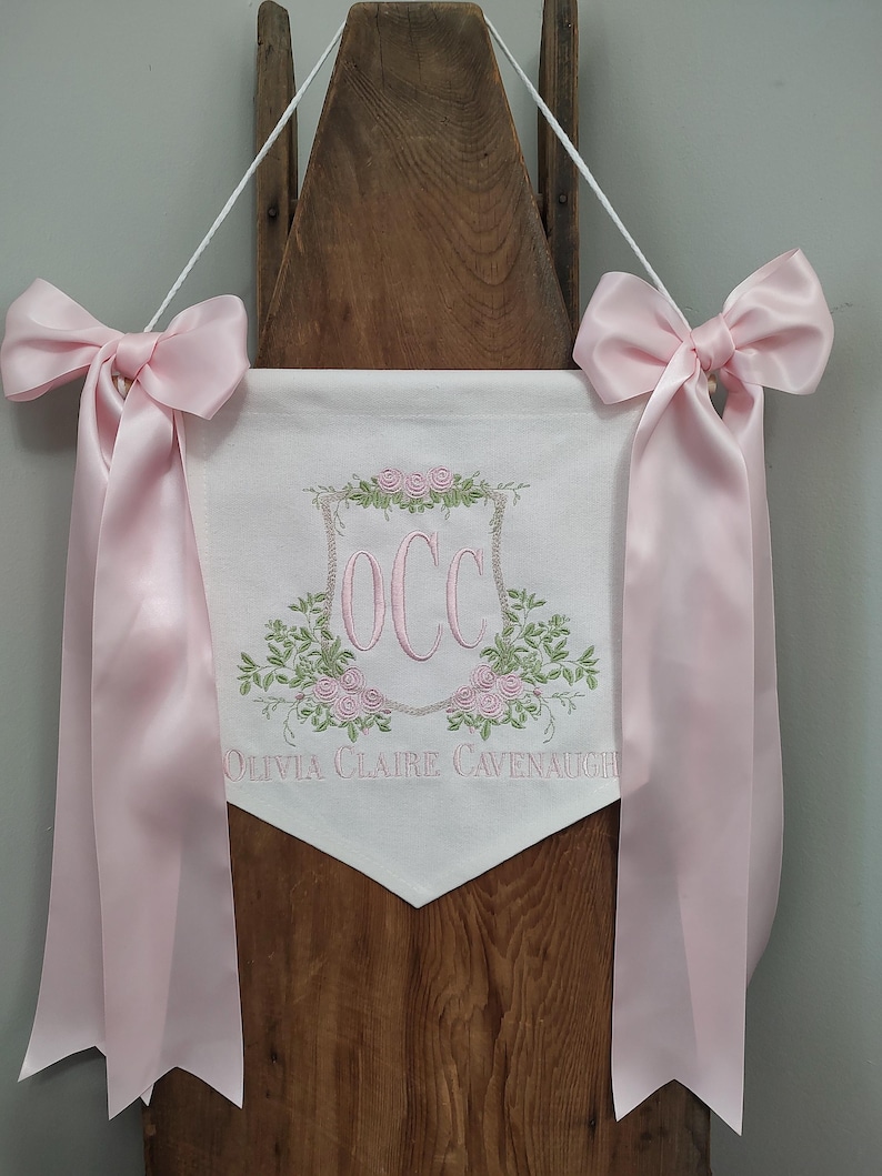 May include: A white banner with pink embroidered floral accents and the initials "OCC" in pink. The banner has the name "Olivia Claire Cavenaugh" embroidered below the initials. Pink satin bows and ribbons are attached to the top.
