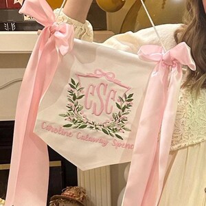 May include: A white banner with pink embroidered lettering and a floral wreath design. The banner has pink ribbons tied at the top corners. The text on the banner reads "CSC" and "Caroline Calaway Spence".