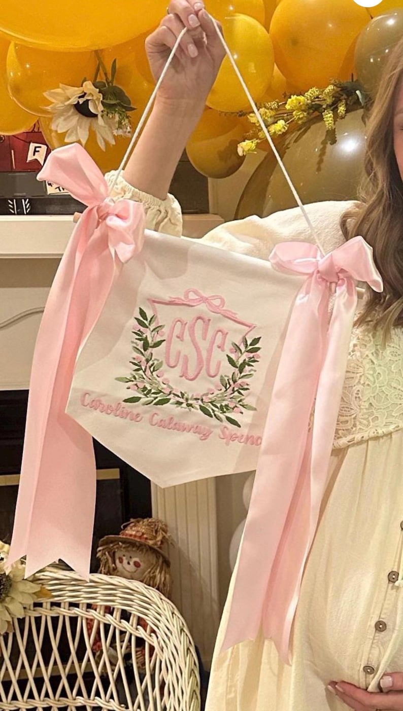 May include: A white banner with pink embroidered lettering and a floral wreath design. The banner has pink ribbons tied at the top corners. The text on the banner reads "CSC" and "Caroline Calaway Spence".