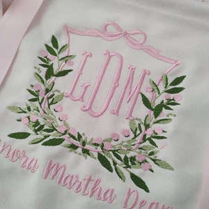 May include: White banner with pink embroidered floral wreath and initials "LDM". The banner has pink ribbon ties and the name "Lenora Martha Dean" embroidered in pink. Green leaves and pink berries complete the wreath design.