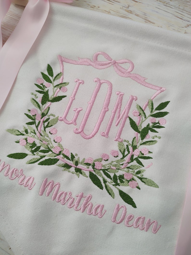 May include: White banner with pink embroidered floral wreath and initials "LDM". The banner has pink ribbon ties and the name "Lenora Martha Dean" embroidered in pink. Green leaves and pink berries complete the wreath design.