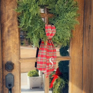 May include: A green wreath with a red and green plaid ribbon tied around it. The ribbon has a white letter "S" on it. The wreath is hanging on a wooden door. The text "Love my wreath sashes made by Miranda! Thank you!!" is at the bottom of the image.