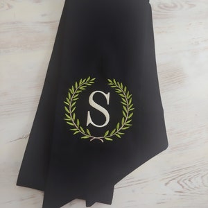 May include: Black graduation sash with a green laurel wreath embroidered with a white letter "S" in the center.