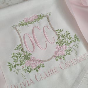 May include: White fabric with pink embroidery featuring the initials "OCC" inside a floral wreath. The name "Olivia Claire Cavenaugh" is embroidered below. A pink satin ribbon is visible.