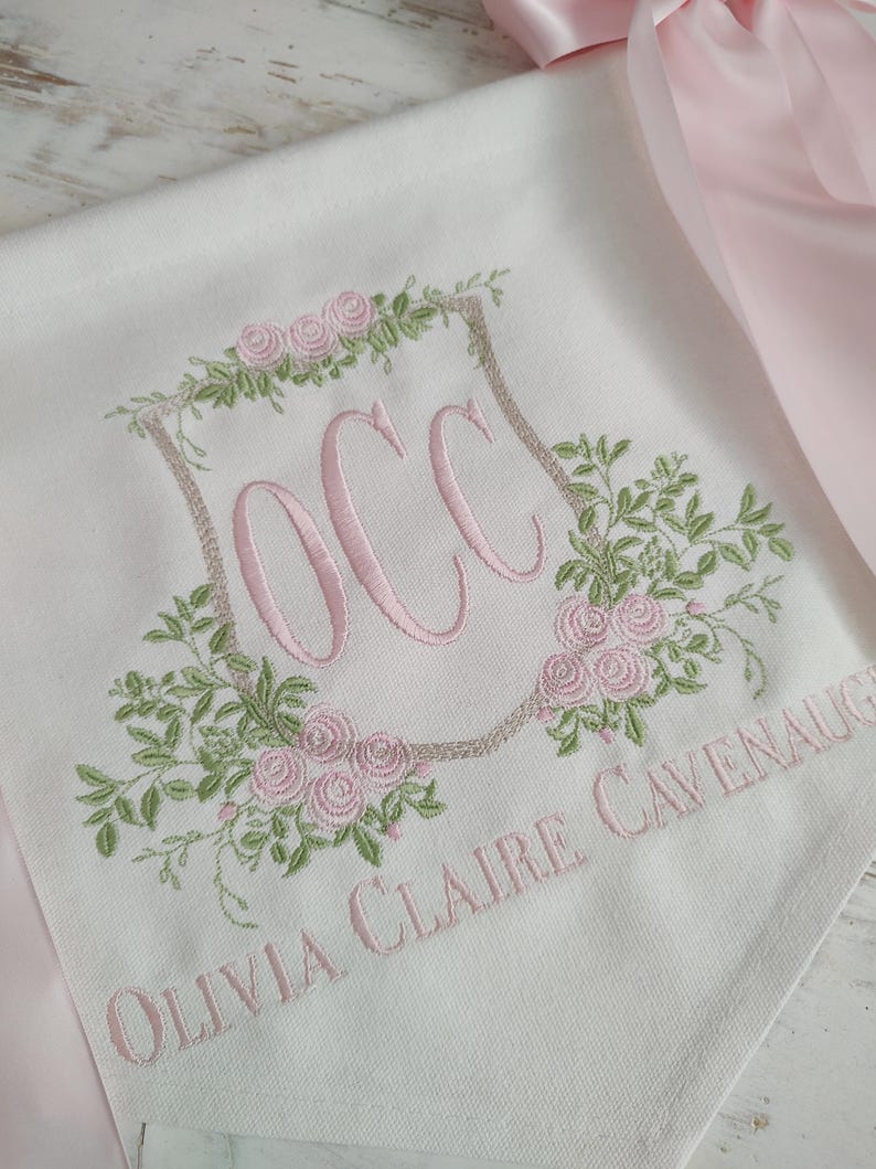 May include: White fabric with pink embroidery featuring the initials "OCC" inside a floral wreath. The name "Olivia Claire Cavenaugh" is embroidered below. A pink satin ribbon is visible.