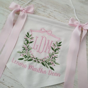 May include: White banner with pink embroidered initials inside a floral wreath. The banner has pink bows and ribbons at the top, and the name "Lenora Martha Dean" is embroidered below the wreath. The banner is hanging from a white string.