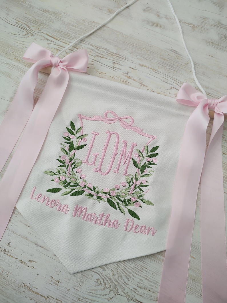 May include: White banner with pink embroidered initials inside a floral wreath. The banner has pink bows and ribbons at the top, and the name "Lenora Martha Dean" is embroidered below the wreath. The banner is hanging from a white string.