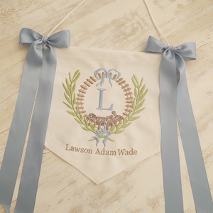 K&ouml;nnte beinhalten: Cremefarbenes Banner mit blauer Stickerei und B&auml;ndern. Das Banner zeigt ein Kranzdesign mit einer Lokomotive und dem Buchstaben "L". Der Name "Lawson Adam Wade" ist darunter gestickt. Zwei blaue Satin-Schleifen schm&uuml;cken die oberen Ecken.