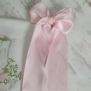May include: A large, light pink satin bow with long, flowing ribbons. The bow is tied in the center and is the focal point of the image. A white cloth with floral embroidery is partially visible.