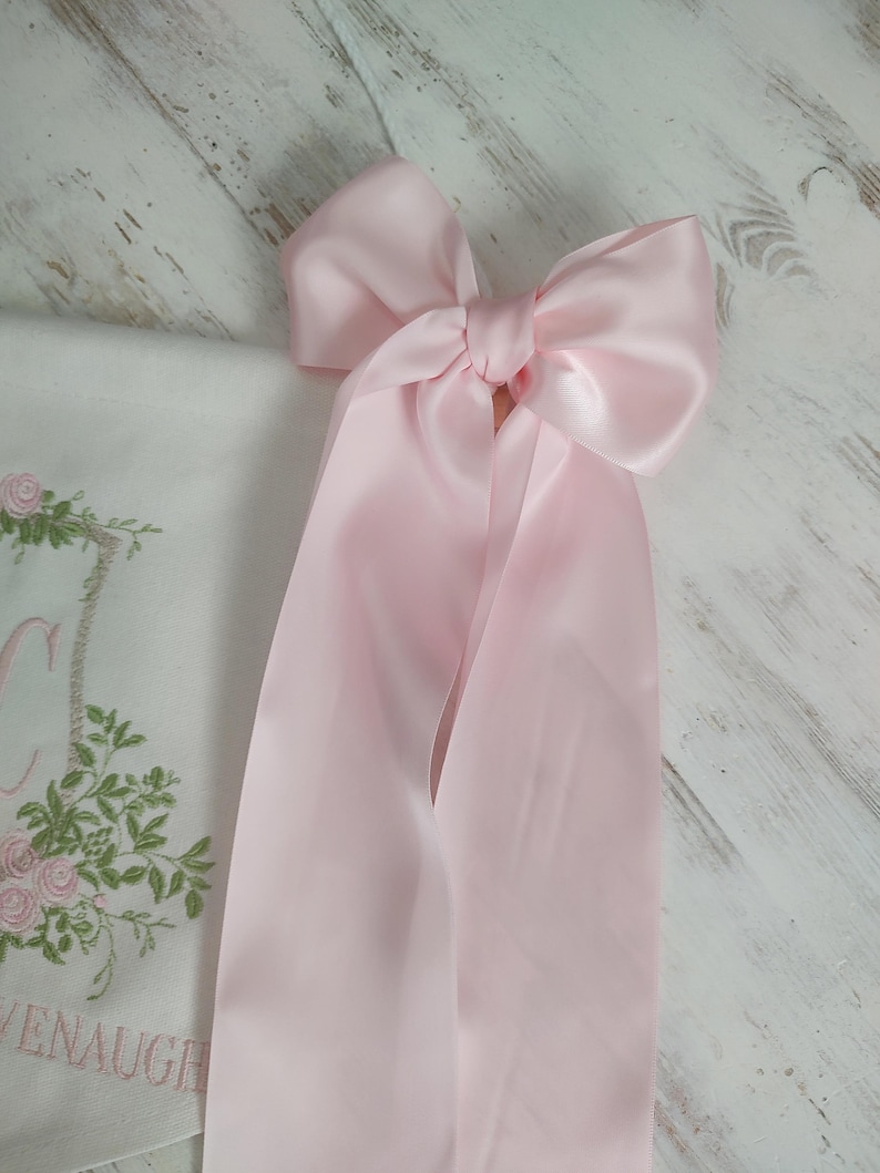 May include: A large, light pink satin bow with long, flowing ribbons. The bow is tied in the center and is the focal point of the image. A white cloth with floral embroidery is partially visible.