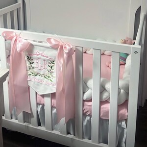 May include: A white crib with pink bedding and a decorative banner. The banner is white with embroidered initials and floral accents, and is tied with pink ribbons. A white braided bumper and a stuffed animal are also visible.