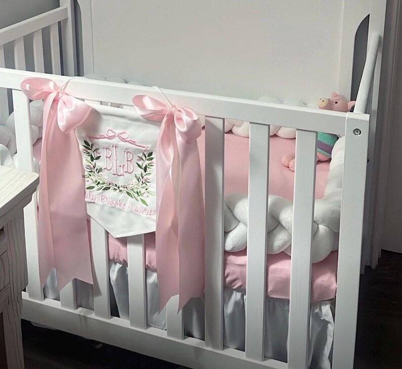 May include: A white crib with pink bedding and a decorative banner. The banner is white with embroidered initials and floral accents, and is tied with pink ribbons. A white braided bumper and a stuffed animal are also visible.