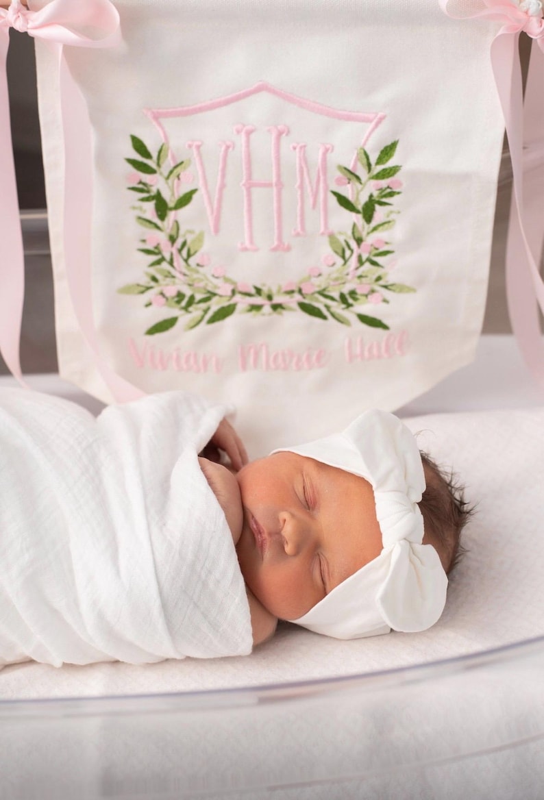 May include: A newborn baby wrapped in a white blanket and wearing a white bow headband. A banner in the background features the embroidered initials "VHM" within a pink shield, surrounded by green leaves and pink flowers, with the name "Vivian Marie Hall" below.