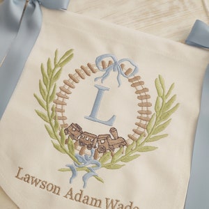 K&ouml;nnte beinhalten: Cremefarbener Banner mit blauen Schleifen. Das Banner zeigt ein gesticktes Design mit einer Lokomotive, einem Kranz und dem Buchstaben "L". Der Name "Lawson Adam Wade" ist darunter gestickt.