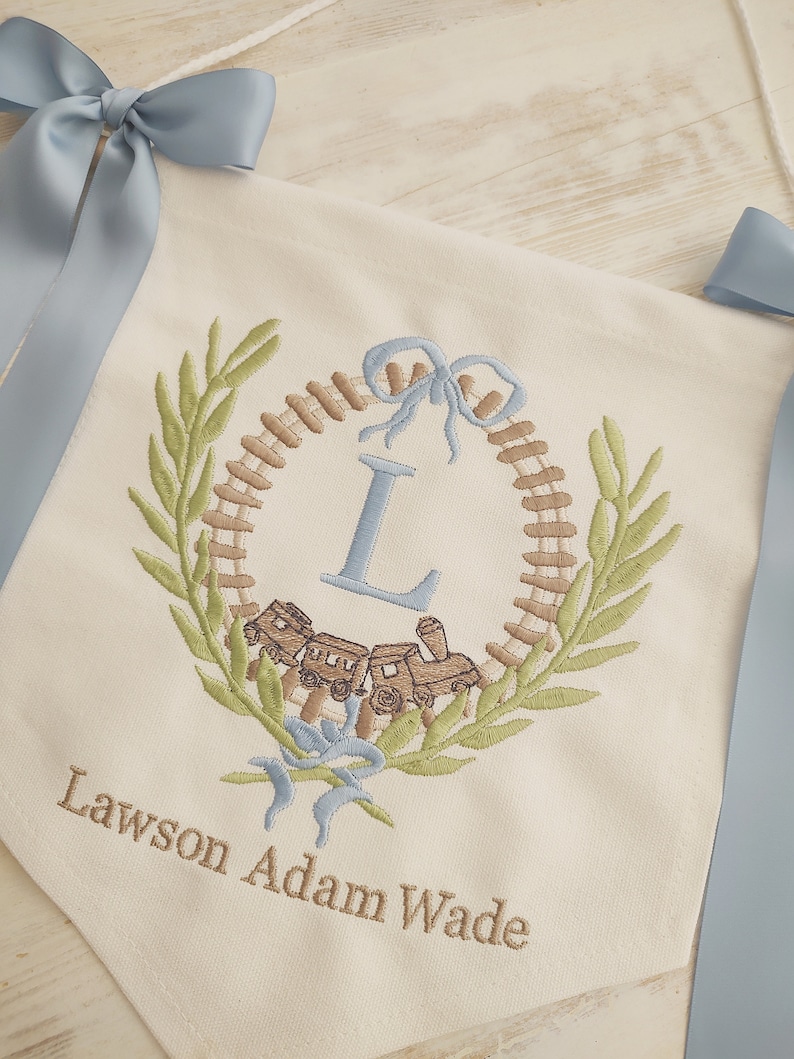 K&ouml;nnte beinhalten: Cremefarbener Banner mit blauen Schleifen. Das Banner zeigt ein gesticktes Design mit einer Lokomotive, einem Kranz und dem Buchstaben "L". Der Name "Lawson Adam Wade" ist darunter gestickt.