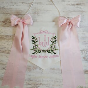 May include: White pennant banner with pink embroidered floral design and the name "Layla James Justice". The banner has pink satin bows and ribbons attached. A white cord is used for hanging.