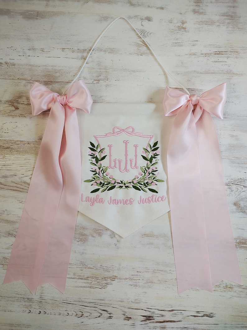 May include: White pennant banner with pink embroidered floral design and the name "Layla James Justice". The banner has pink satin bows and ribbons attached. A white cord is used for hanging.