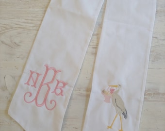 May include: White fabric sash with pink embroidered monogram "NRB" and a pink and yellow embroidered stork carrying a baby in a pink blanket.