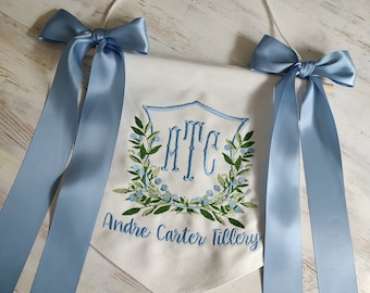 May include: A white banner with a light blue embroidered design, including initials and a name, is adorned with light blue satin bows. The banner is suspended by a white cord and is suitable for nursery decor.