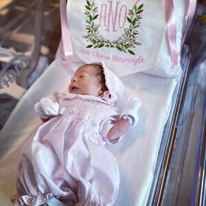 May include: A newborn baby wearing a light pink gown and bonnet, lying on a white surface. A decorative pillow with the initials "ANO" and the name "Ava Olivia Normoyle" is visible in the background. The pillow features a floral wreath design.