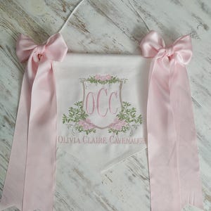 May include: A personalized banner with the initials "OCC" embroidered in pink, surrounded by floral designs. The banner includes the name "Olivia Claire Cavenaugh" and is adorned with pink bows and ribbons.