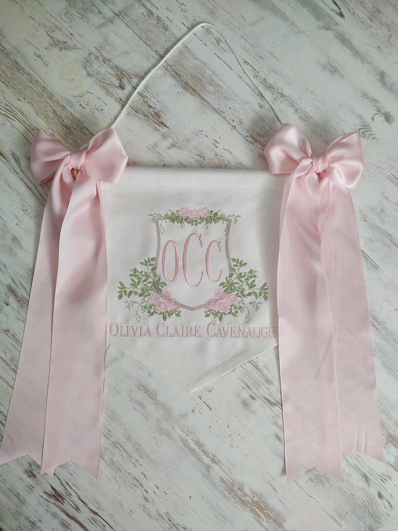 May include: A personalized banner with the initials "OCC" embroidered in pink, surrounded by floral designs. The banner includes the name "Olivia Claire Cavenaugh" and is adorned with pink bows and ribbons.