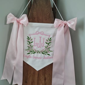 May include: White banner with pink embroidered details and pink satin ribbons. The banner features a floral wreath design with the initials "LJJ" and the name "Layla James Justice". The banner is suspended by a white cord.