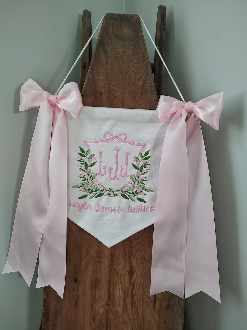 May include: White banner with pink embroidered details and pink satin ribbons. The banner features a floral wreath design with the initials "LJJ" and the name "Layla James Justice". The banner is suspended by a white cord.