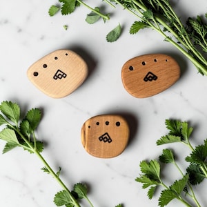 May include: Three wooden soap dishes in varying shades of brown and tan, each with a unique shape and a carved design. The dishes have small holes and a logo. Fresh green herbs surround the dishes on a white marble surface.