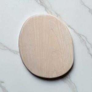 May include: A light-colored, oval-shaped wooden serving board with a natural wood grain pattern. The board has a smooth surface and rounded edges. The board is set against a white marble background.